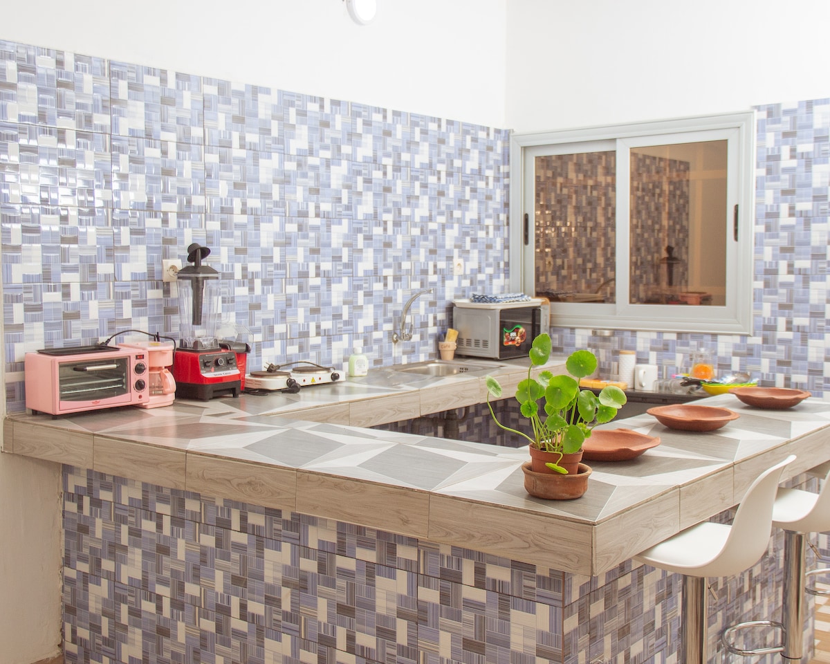 A well-equipped kitchen features tiled walls in a mosaic pattern. Countertops hold various appliances, including a pink toaster and a stove. A potted plant adds a touch of greenery. A large window allows natural light to fill the space, enhancing the inviting atmosphere.