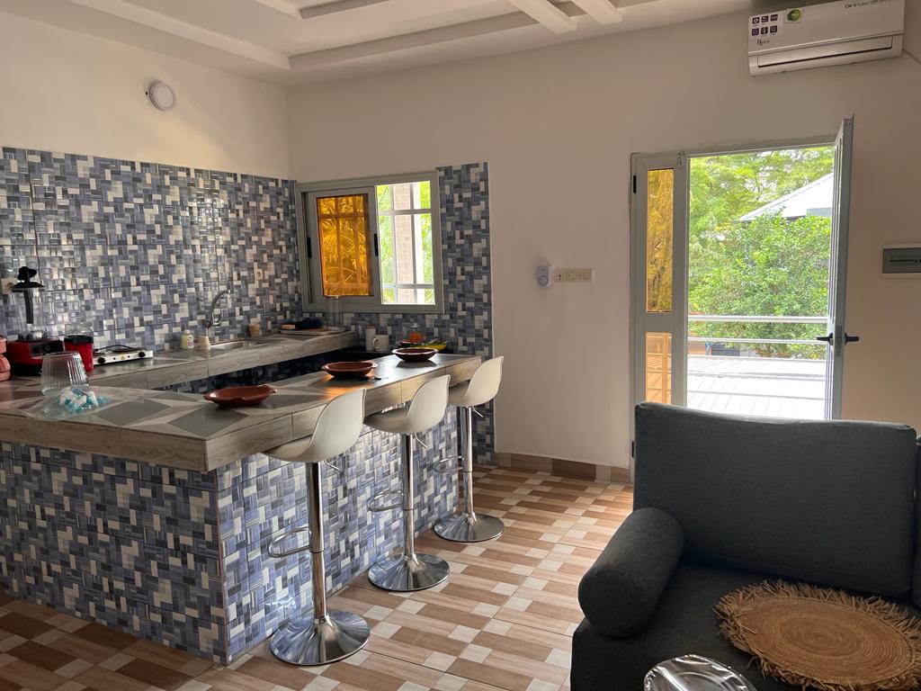 A modern kitchen area features a patterned tile backsplash and a breakfast bar with four white stools. Large windows allow natural light to fill the space, and doors lead to an outdoor area surrounded by greenery. A gray sofa adds comfort to the setting.