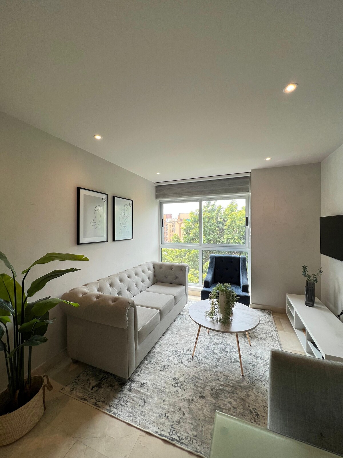 A comfortable seating area features a light-colored couch and a dark armchair, arranged around a round coffee table. Natural light enters through a large window, illuminating the plants and decorative elements within the space.