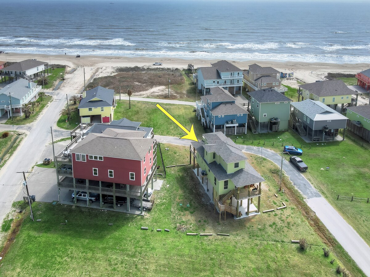An aerial view showcases the two-story house in relation to the nearby beach. The proximity of the home to the shoreline is highlighted, along with surrounding houses and open grassy areas, creating a sense of space and accessibility.