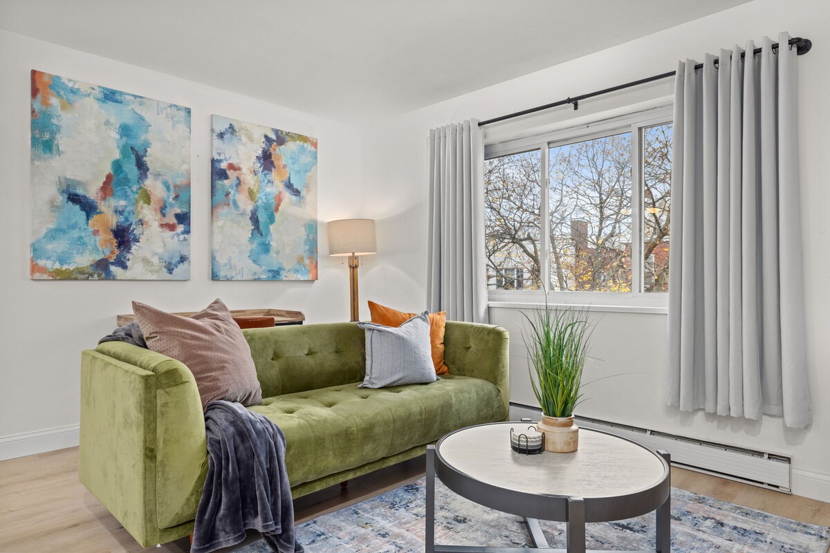 A cozy living area features a green sofa adorned with decorative pillows and a soft throw blanket. A round coffee table sits in front, complemented by a small plant. Two colorful paintings enhance the space, while natural light filters through a window dressed with light curtains.