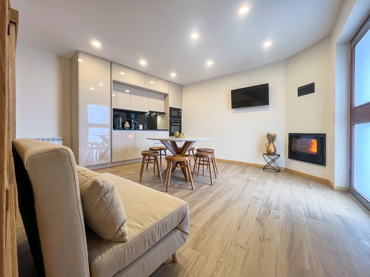 An open-plan living area features a modern kitchen with sleek cabinetry and a dining table surrounded by four wooden chairs. A cozy fireplace is positioned nearby, with a television mounted on the wall. Natural light fills the space through large windows.
