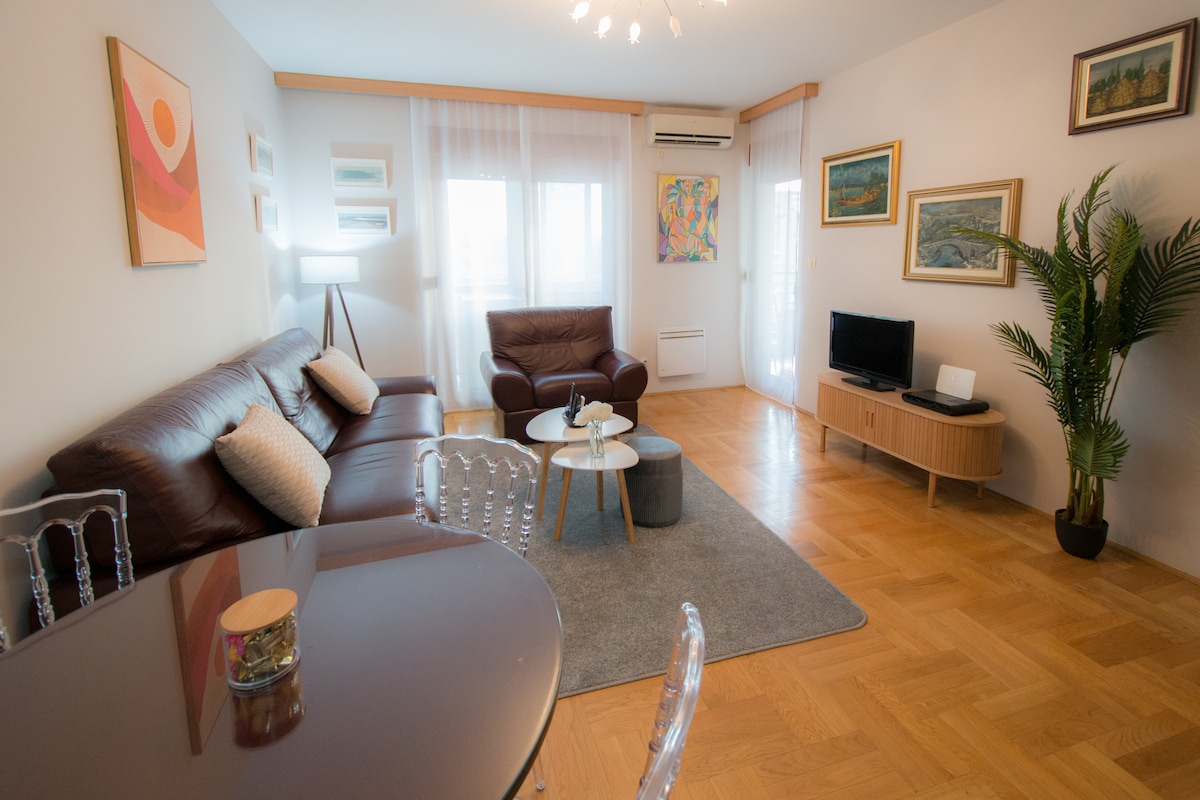 A spacious living area is presented, featuring a dark leather sofa and armchair complemented by a light-colored rug. Art pieces adorn the walls, and a television is positioned on a wooden console. Natural light filters in through sheer curtains, creating an inviting atmosphere.