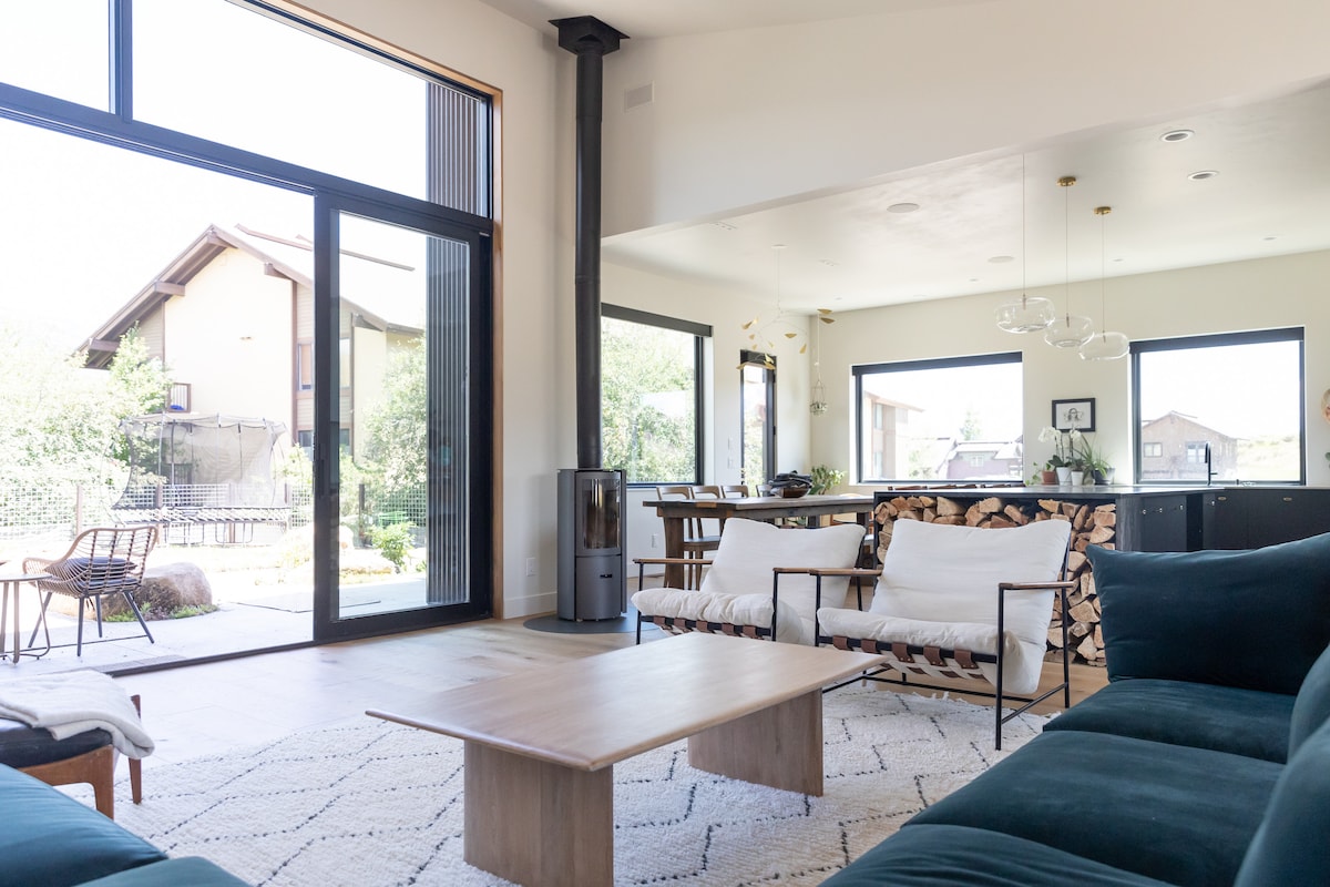 A bright, open living space features large glass doors that connect to the outdoor area. Comfortable seating is arranged around a low wooden table, with a light rug beneath. A contemporary dining area is visible in the background, accentuated by natural light.