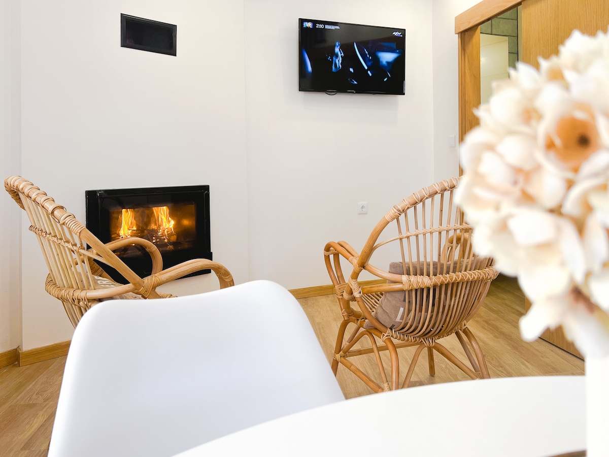 A cozy sitting area features two woven chairs positioned beside a modern fireplace. A wall-mounted television is visible, while a dining table with a minimalist design provides a casual dining space. Natural light enhances the warm tones of the wooden flooring.