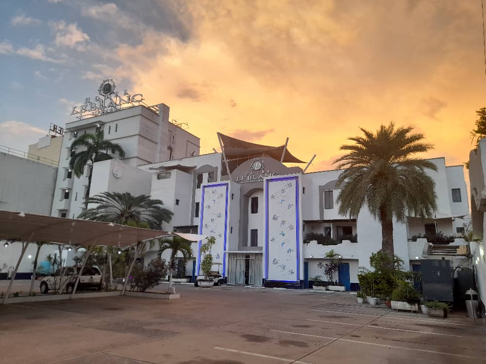 A modern building with a distinctive architectural design is set against a vibrant sunset sky. Palm trees and landscaped plants are arranged in the foreground, enhancing the welcoming appearance of the entrance. Covered parking spaces are visible to the side.