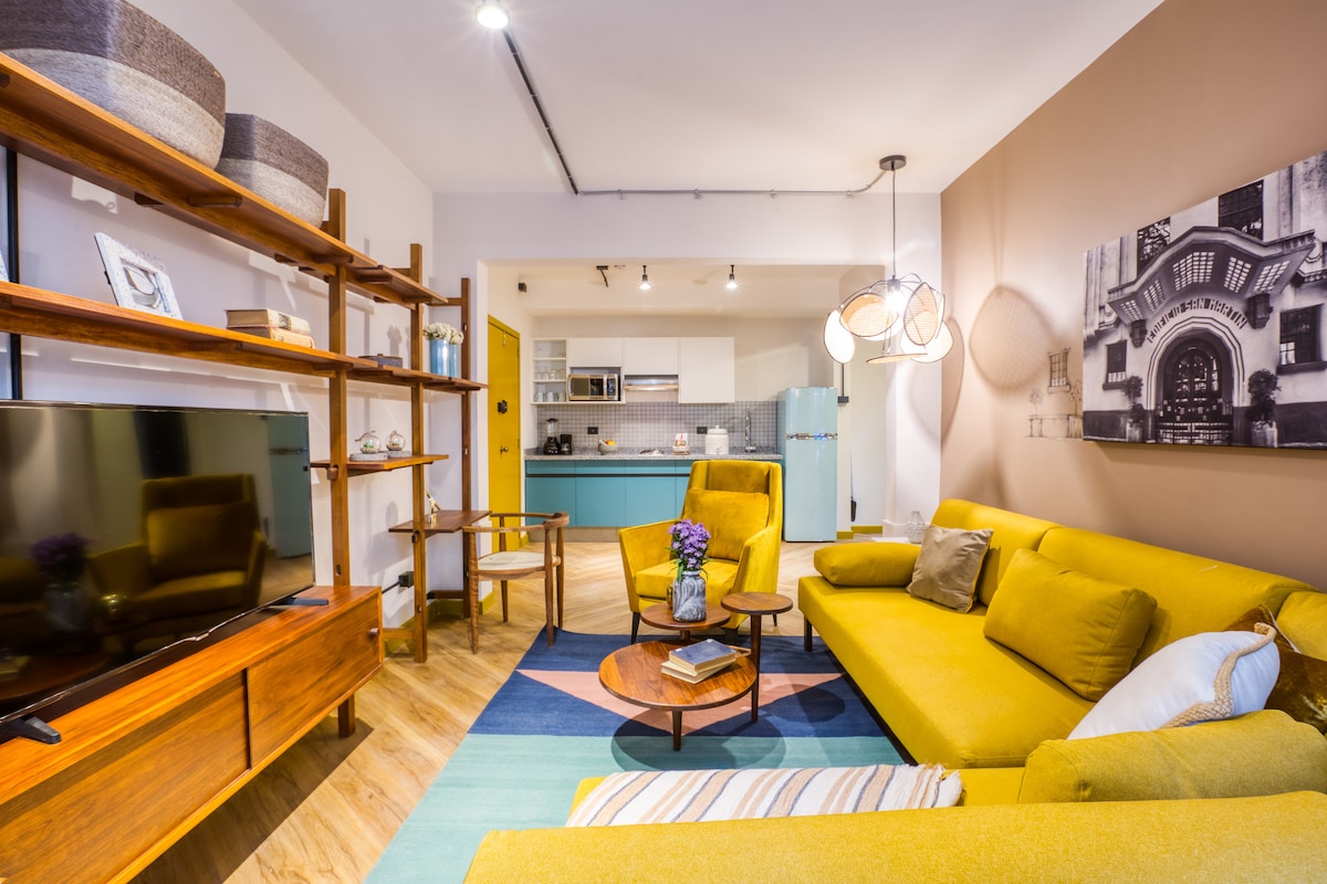 A cozy living area features a bright yellow sofa complemented by wooden side tables. A large television is mounted on a wooden shelf, adorned with decorative items. The space includes a multi-colored rug, with an open view leading to a modern kitchen area.