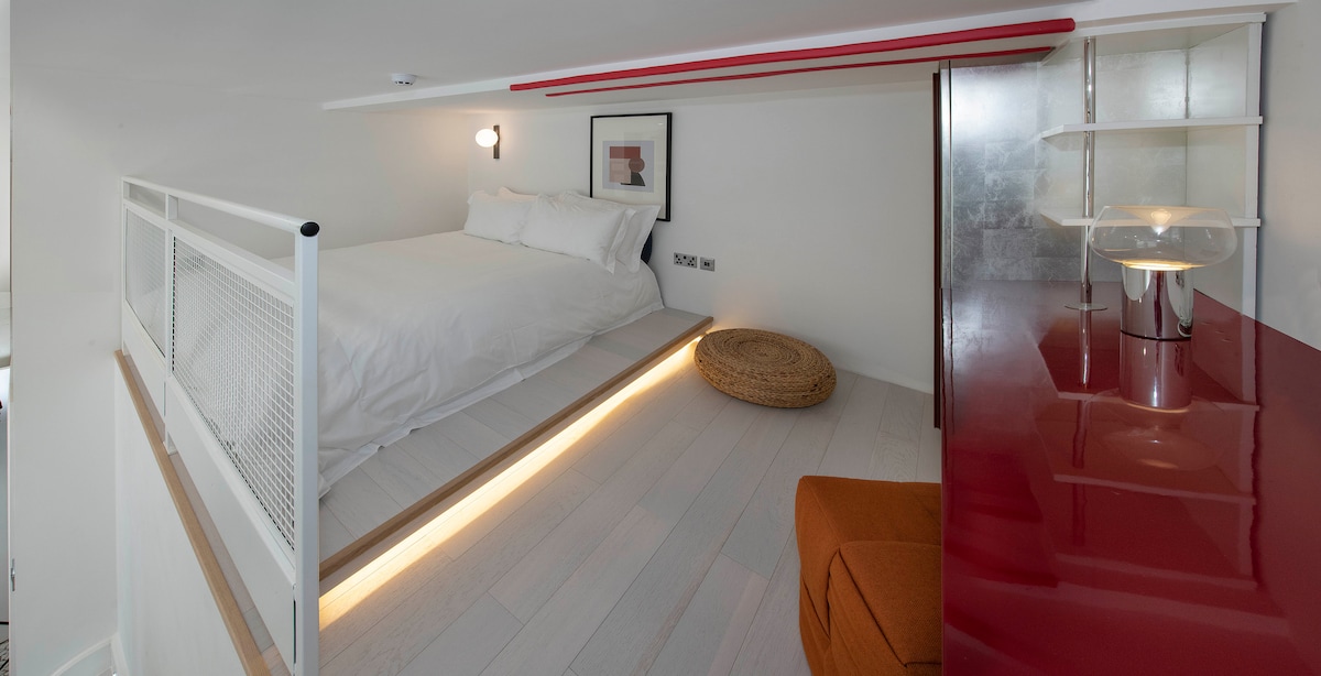 A compact sleeping area is located on a raised platform, featuring a spacious bed with crisp white linens. Soft lighting highlights the minimalist design, while a warm-toned rug adds texture beneath. Nearby, a sleek red accent table and a modern lamp contribute to the overall aesthetic.