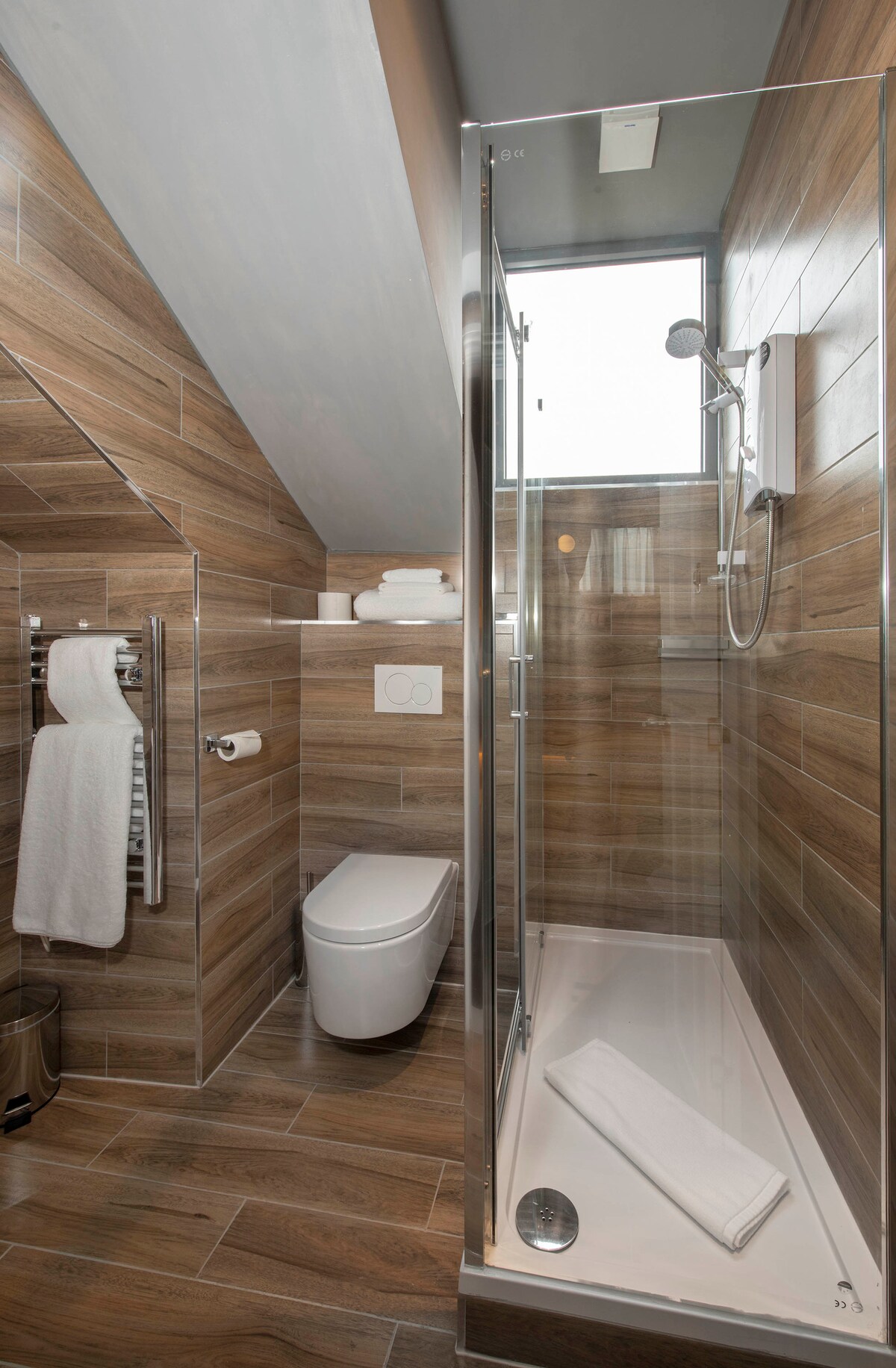 A modern bathroom is showcased with wood-toned tiles enhancing warmth. A glass shower enclosure is visible, featuring a rain shower head. Soft towels are neatly arranged above the toilet, and a sleek toilet complements the design. Natural light streams in from a nearby window.