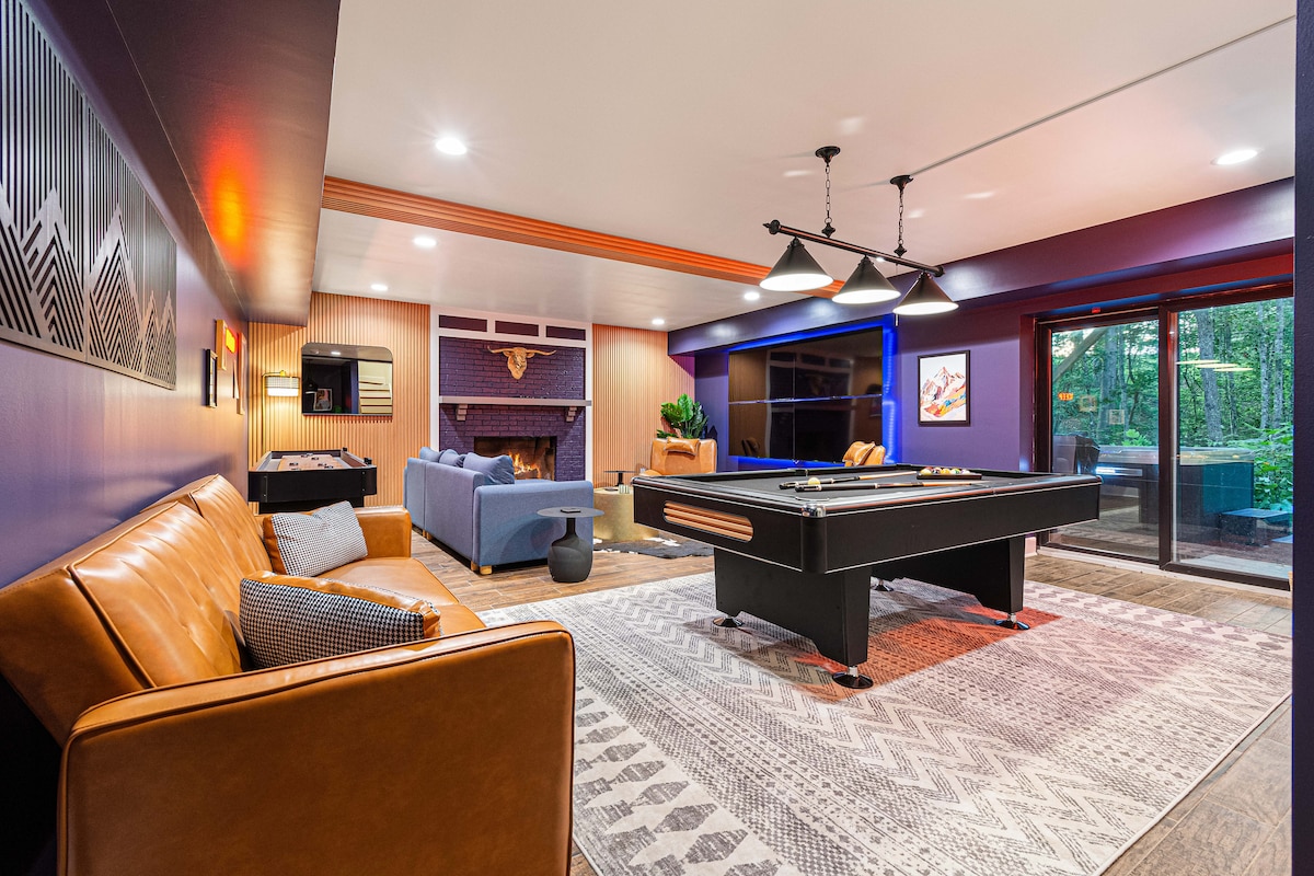 A cozy game room features a pool table as its centerpiece, accompanied by a leather couch and modern seating. Warm lighting highlights the room's design, while large glass doors provide a view of the outdoor space. A fireplace adds a welcoming touch.