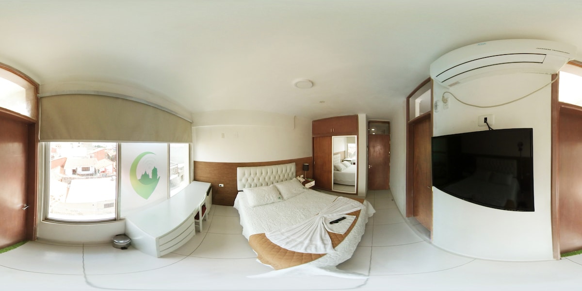 A bright and airy bedroom features a spacious bed with white linens. A large window allows natural light to fill the room, while a flat-screen TV is mounted on the wall. A mirror and a small desk add convenience in a minimalistic design.