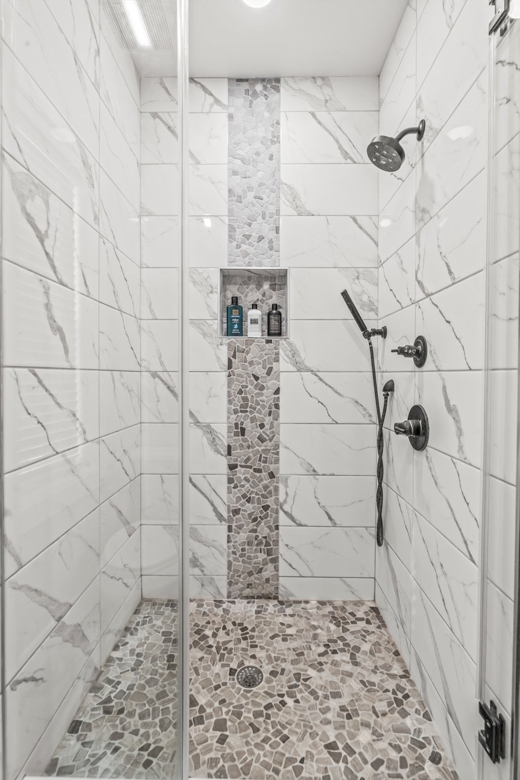 A modern shower stall features sleek white tiles with gray veining and a decorative stone inlay. The spacious shower includes a rainfall showerhead and a handheld shower attachment. Shelves hold various toiletries, and the flooring consists of small, textured stones.