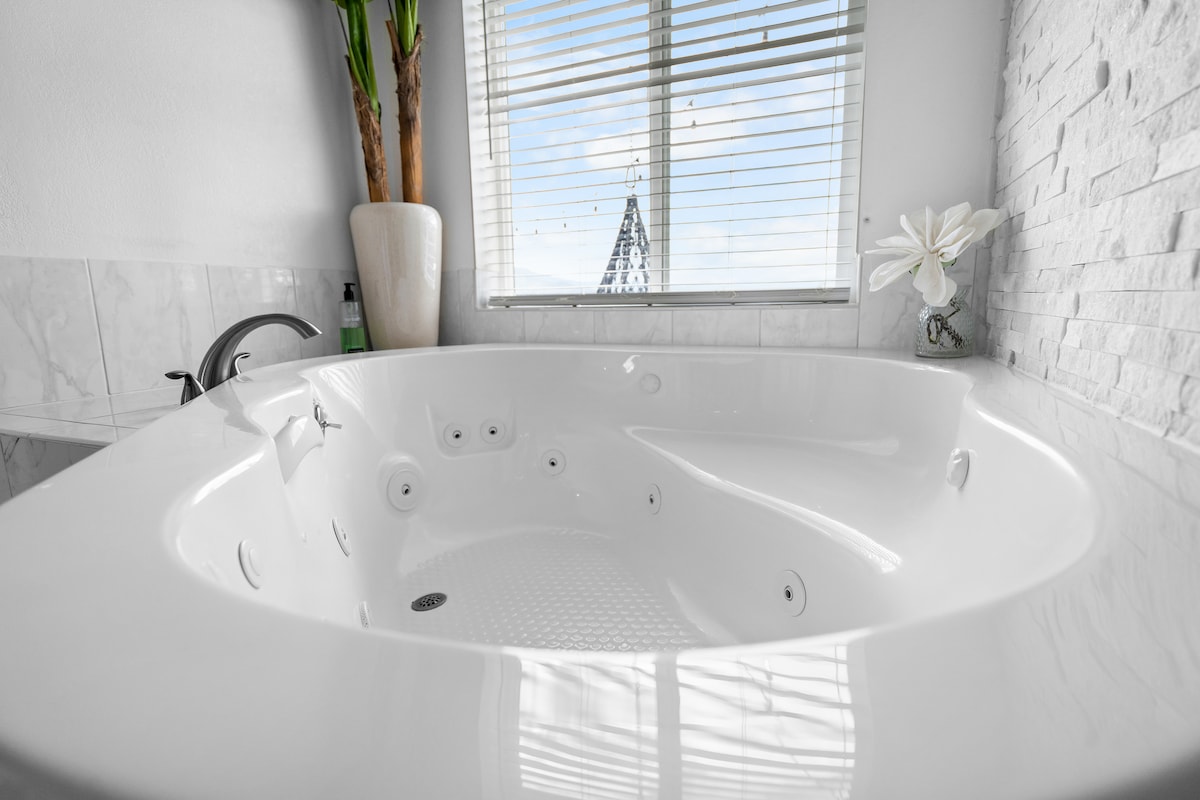 A jetted tub is prominently featured, surrounded by elegant stone walls. Natural light filters through a nearby window with blinds, highlighting the tub's smooth contours. A decorative vase with a flower adds a touch of charm to the serene bathroom environment.