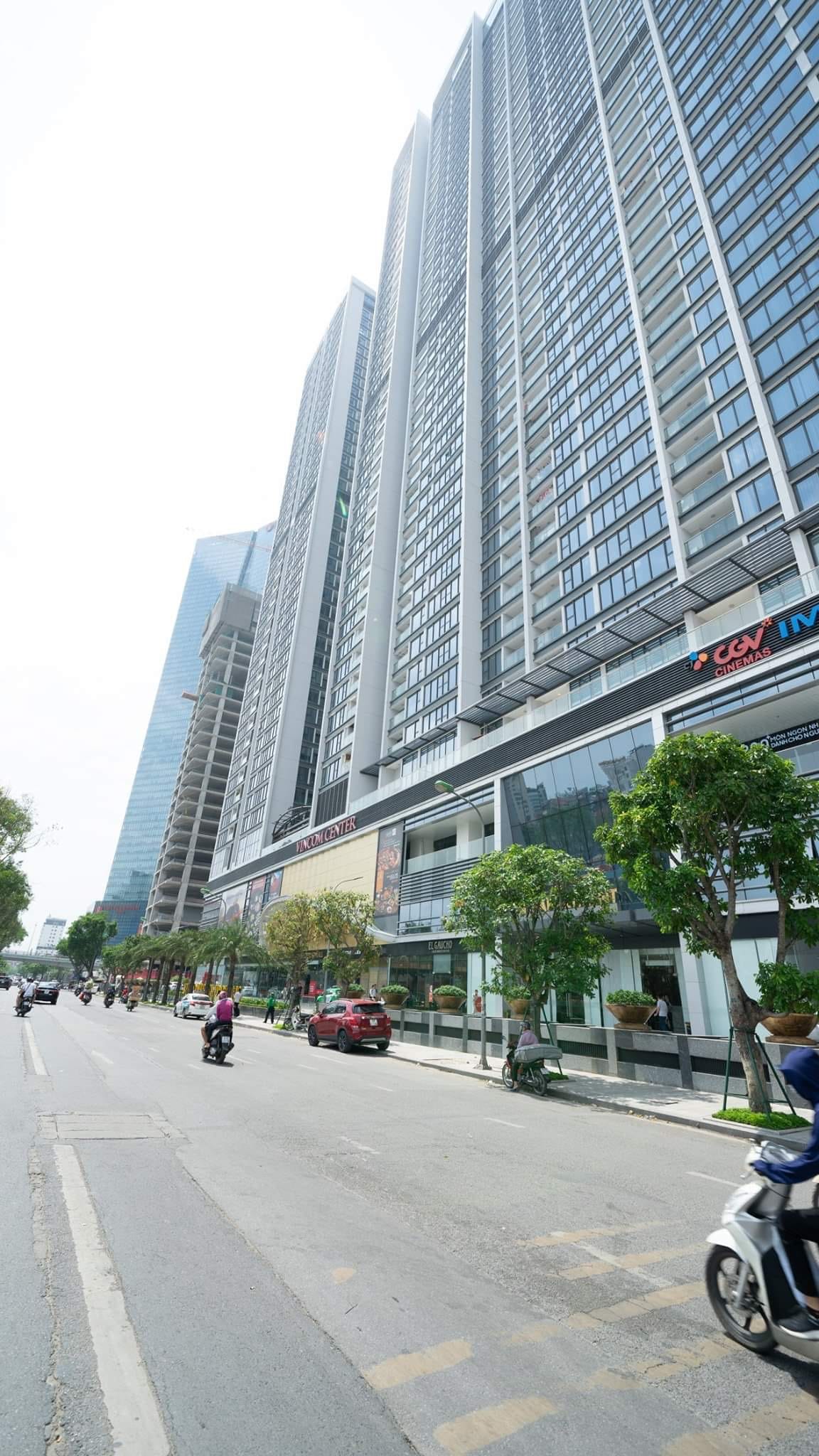 The modern apartment building is viewed from the street, showcasing its tall, glass-fronted facade. Lush greenery, including small trees and shrubs, lines the exterior, while vehicles navigate the nearby road. Pedestrians can be seen enjoying the vibrant urban environment.