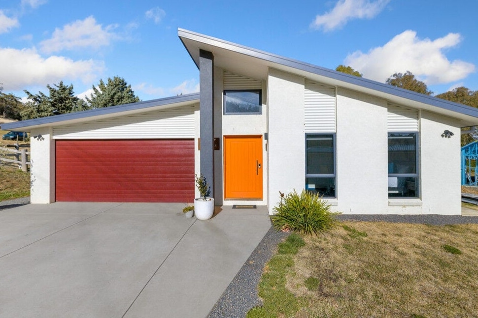 The exterior of a modern home is presented, featuring a striking orange front door against a white facade. A red garage door complements the design. Lush greenery and neatly trimmed grass are visible in the front yard, with a clear blue sky above.