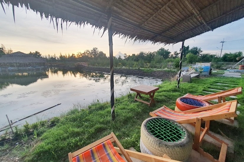 A shaded seating area is positioned beside a calm water body, featuring colorful lounge chairs and rustic wooden tables. The landscape is enhanced by greenery, with a low horizon blending into the early evening sky.