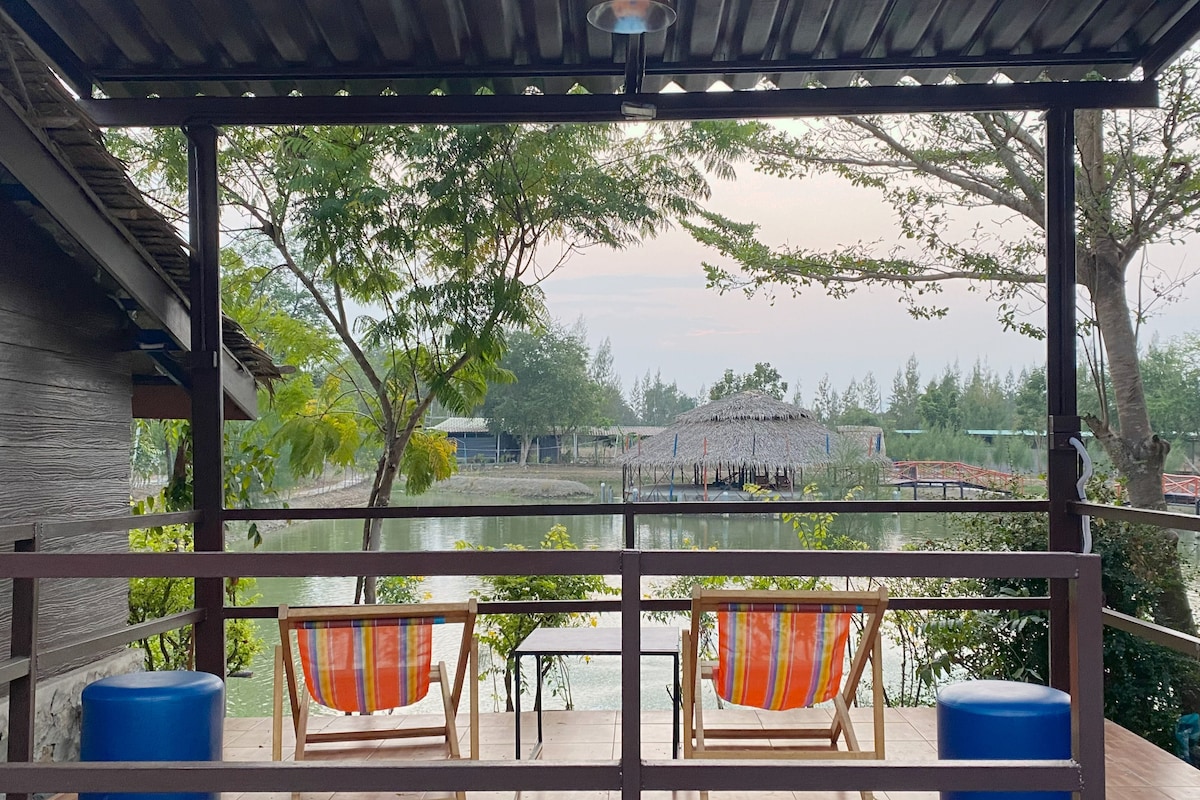A wooden deck features two lounge chairs with colorful striped cushions, facing a serene body of water. A table is positioned between the chairs, and lush greenery surrounds the scene. In the distance, a thatched-roof building is partially visible, framed by trees.
