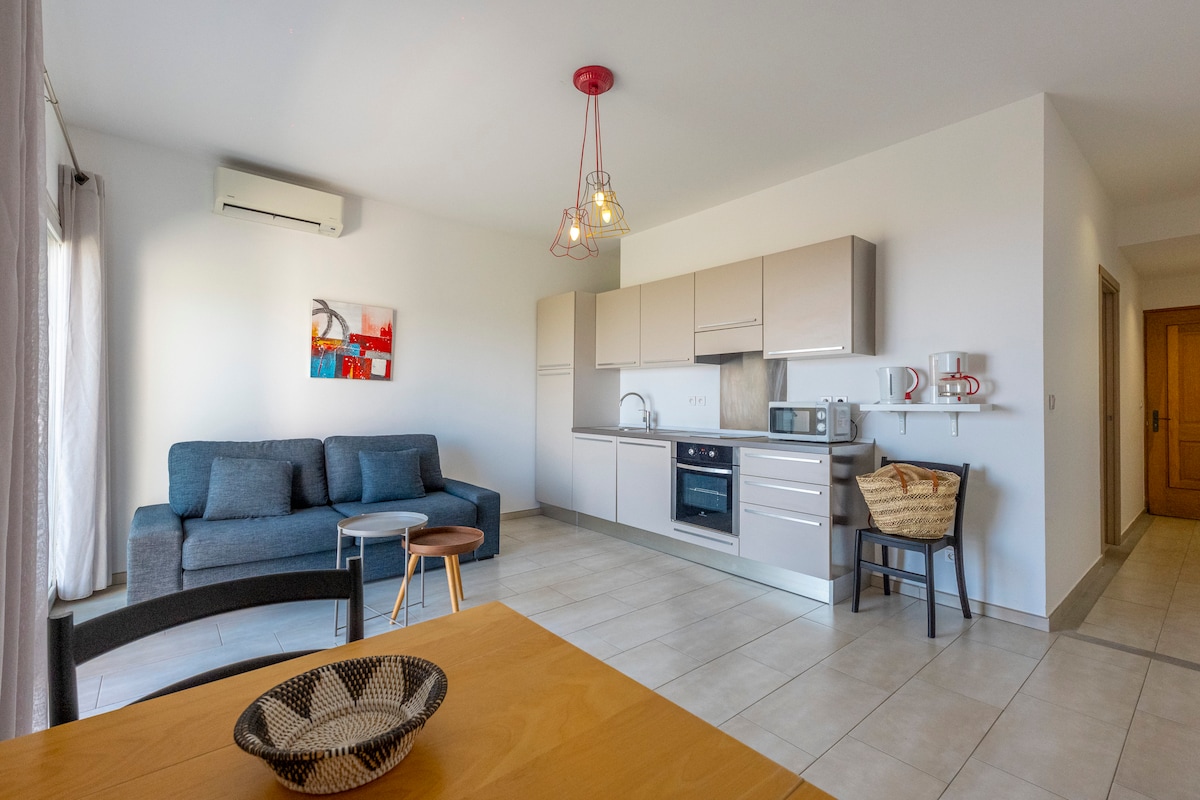 A bright living space is featured, showcasing a comfortable sofa and a small side table. The kitchen area contains modern appliances, including a microwave and stovetop. Light-colored cabinetry complements the neutral tile flooring, while a simple artwork adds a touch of color to the wall.