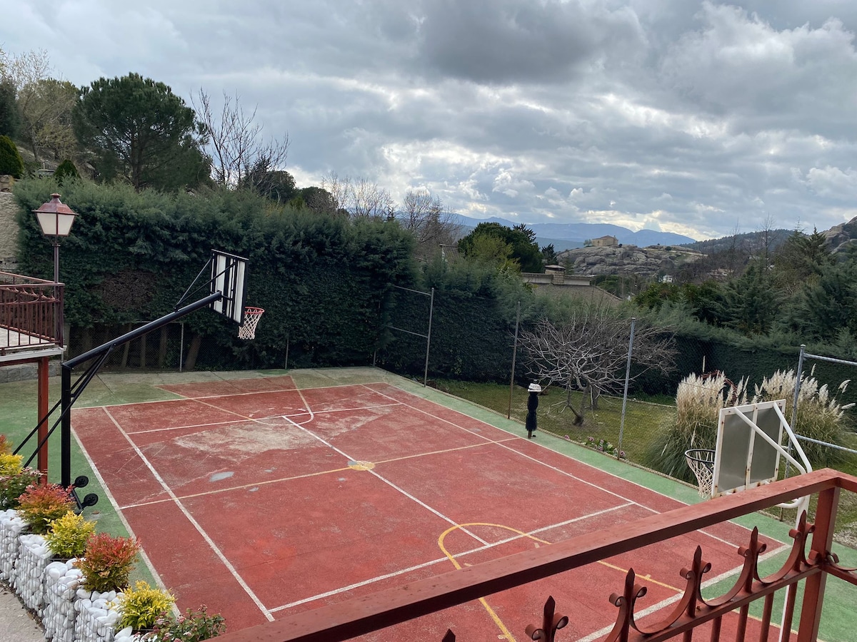 An outdoor basketball court is visible, featuring a well-maintained hoop and a textured playing surface. Lush greenery surrounds the area, with distant hills partially visible in the background. The court is enclosed, providing a private space for recreation.