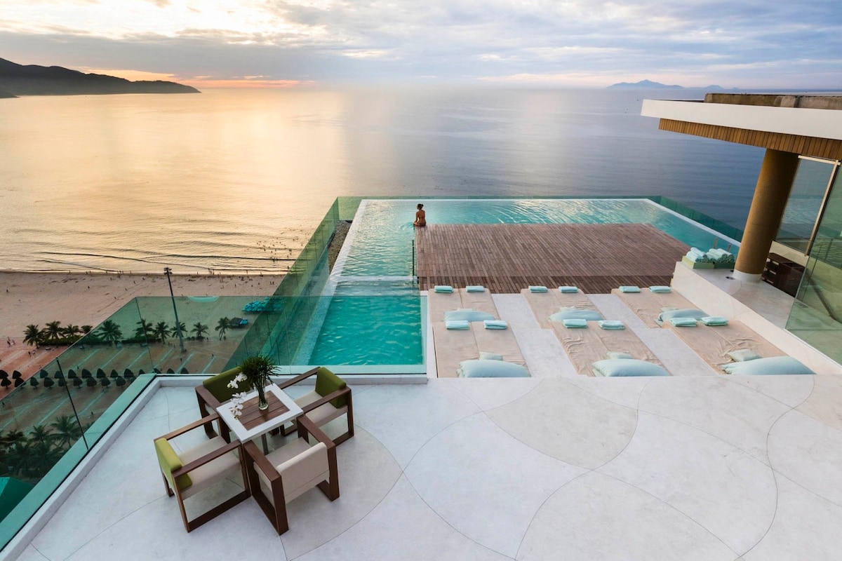 An expansive rooftop terrace features an infinity pool overlooking the calm sea at sunset. Lounge chairs are positioned around the pool, offering a serene space to relax. The view encompasses the shoreline and distant mountains, emphasizing the tranquil setting.