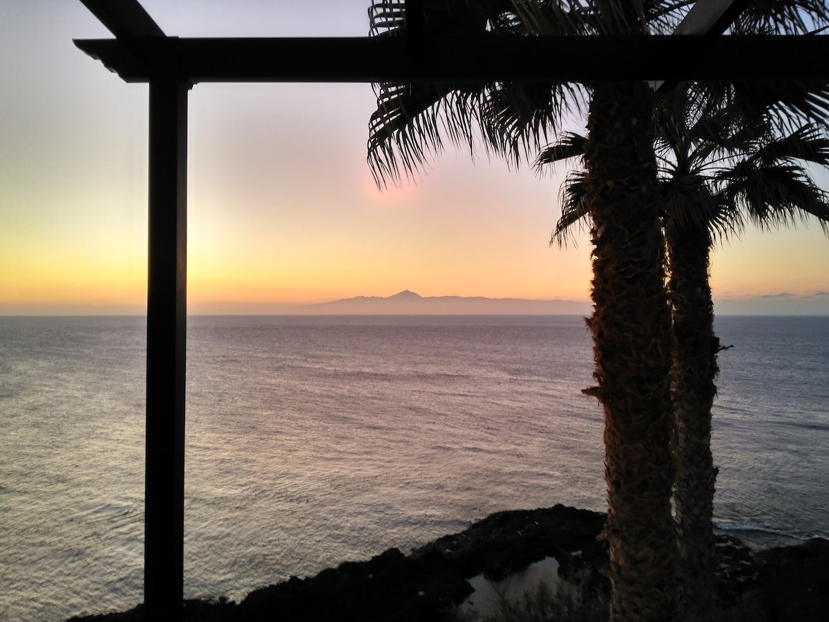 The view captures a serene coastline at sunset, with soft hues of orange and pink illuminating the sky. In the foreground, palm trees frame the scene, and the tranquil ocean reflects the colorful sky, creating a peaceful atmosphere.