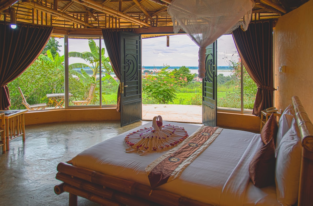A cozy sleeping area features a spacious bed adorned with decorative elements. Large wooden doors open to reveal a view of lush greenery and the Mekong River beyond. Natural light floods the room, enhancing the warm colors of the bamboo design.