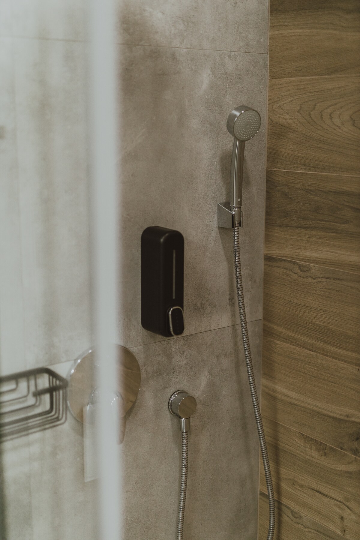 A modern shower area is visible with sleek, minimalist fixtures. A handheld showerhead and a mounted showerhead are installed on a textured gray wall. The design incorporates natural elements with wood accents adding warmth to the space.