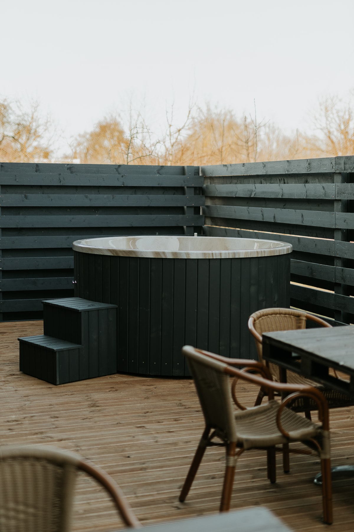 A wooden terrace features a round hot tub surrounded by a wooden deck. Two steps lead up to the tub, while a dining table with wicker chairs is positioned nearby. The backdrop consists of dark wooden fencing, providing a sense of privacy.