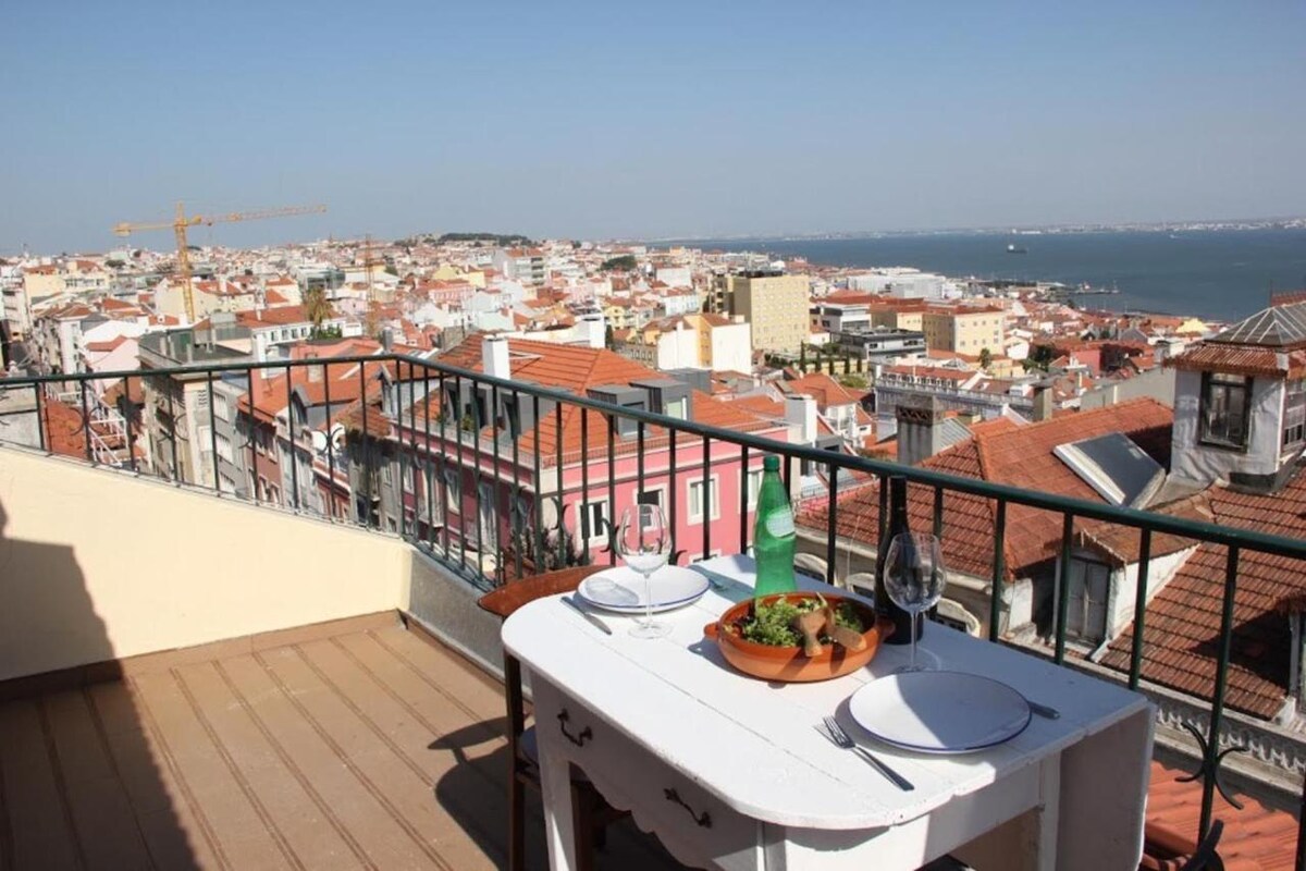 A dining table is set on a sunny terrace, featuring two plates and a glass, overlooking a panoramic view of the city and the river. Brightly colored rooftops are visible in the distance, while a gentle breeze can be felt in the open space.