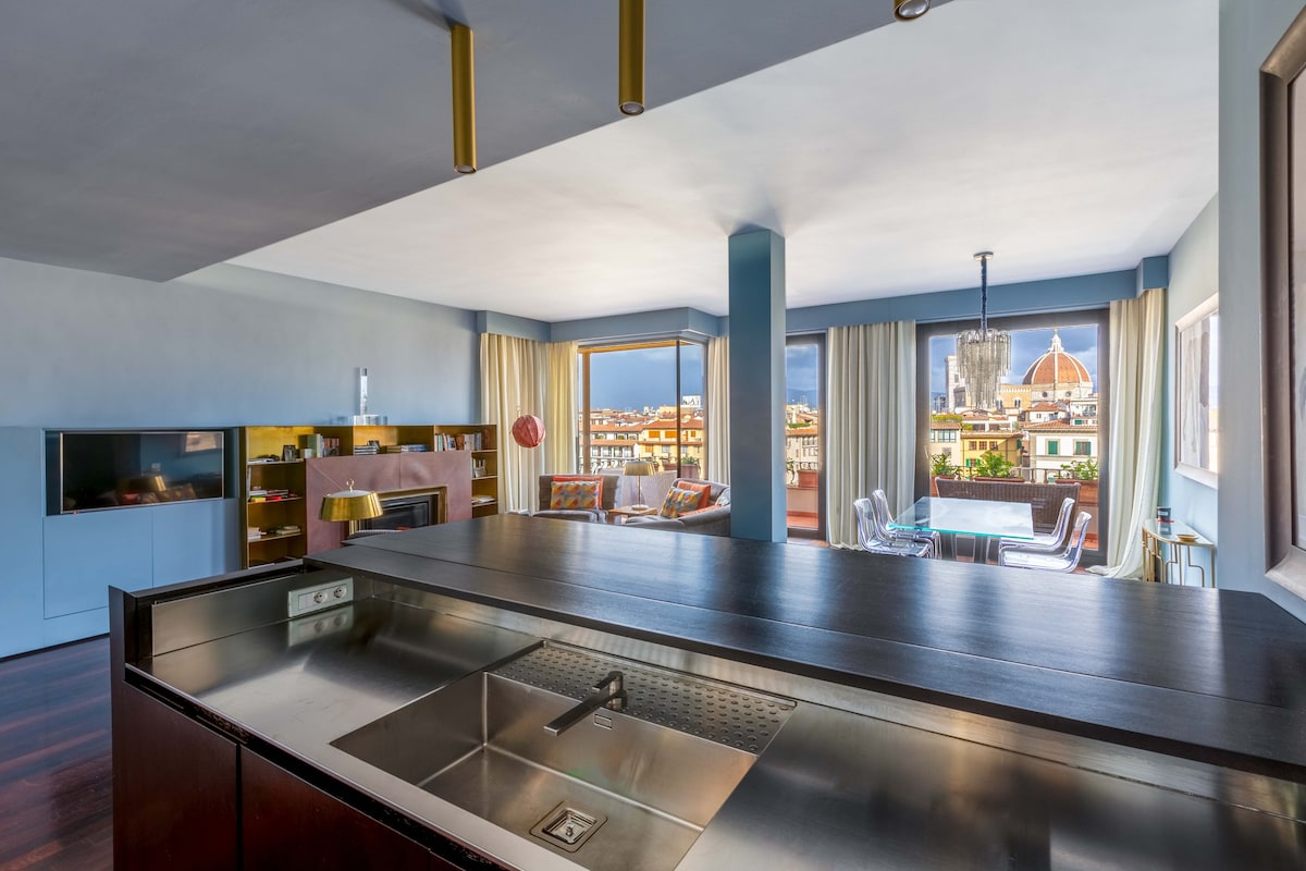 A modern open living space is captured, featuring a large kitchen island with a sink and a view overlooking the city. Large windows provide abundant natural light, revealing views of Florence's skyline with its renowned monuments in the background.