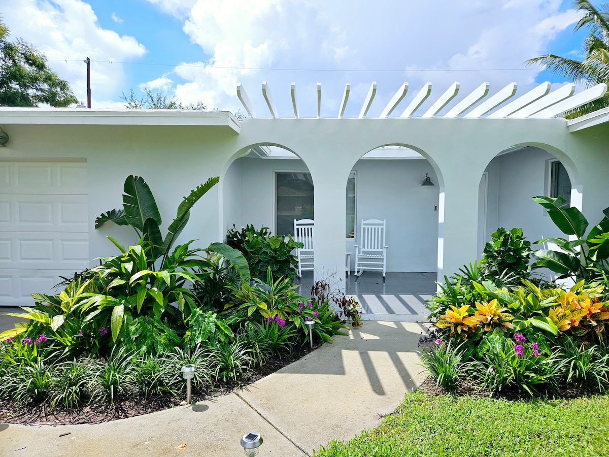 The exterior features a welcoming entryway with two rocking chairs under an architectural archway. Lush greenery and colorful plants adorn the pathway, enhancing the inviting atmosphere of the home. A clean, white facade complements the surrounding nature.