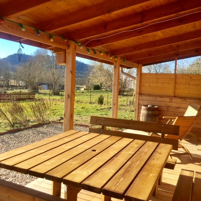 A covered terrace features a large wooden table surrounded by benches. The area is brightened by string lights overhead, and the surrounding landscape includes grassy space and distant mountains, creating a calm outdoor setting.