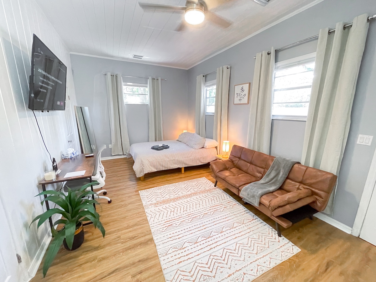 A modern and airy guest room features a spacious bed with soft bedding and two windows allowing natural light. A workspace with a desk and chair is positioned nearby, along with a cozy seating area that includes a leather sofa. A decorative area rug adds warmth to the wooden floor.