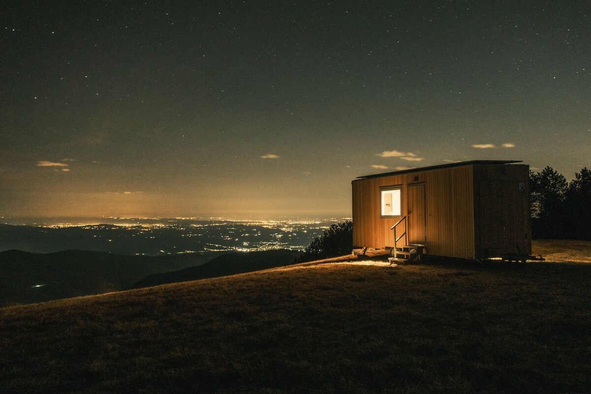 A tiny house is positioned on a hilltop, illuminated against a starry night sky. Soft light emanates from its window, highlighting the surrounding landscape. The view extends over a valley blanketed in soft lights from distant towns, with dark silhouettes of mountains rising in the background.