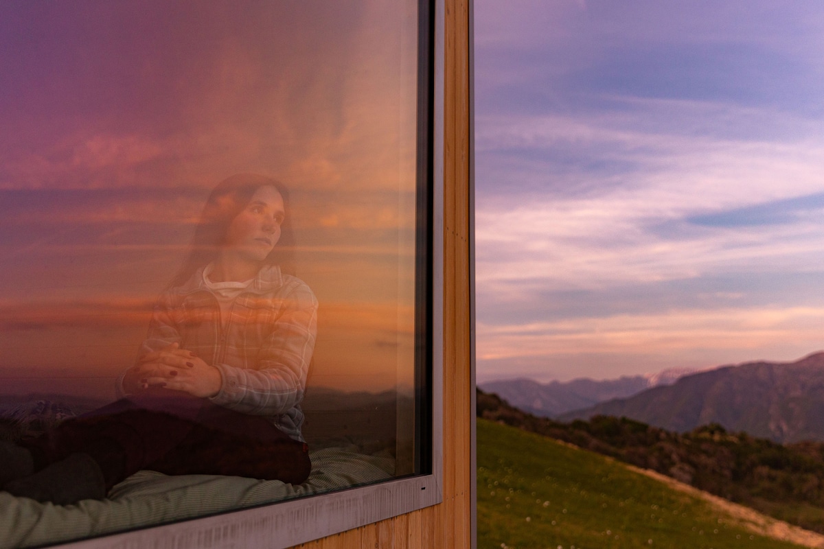 A reflective surface showcases a serene sunset view over rolling hills and distant mountains. The colors transition from warm oranges to cool purples in the sky, blending with the gentle green of the surrounding landscape.