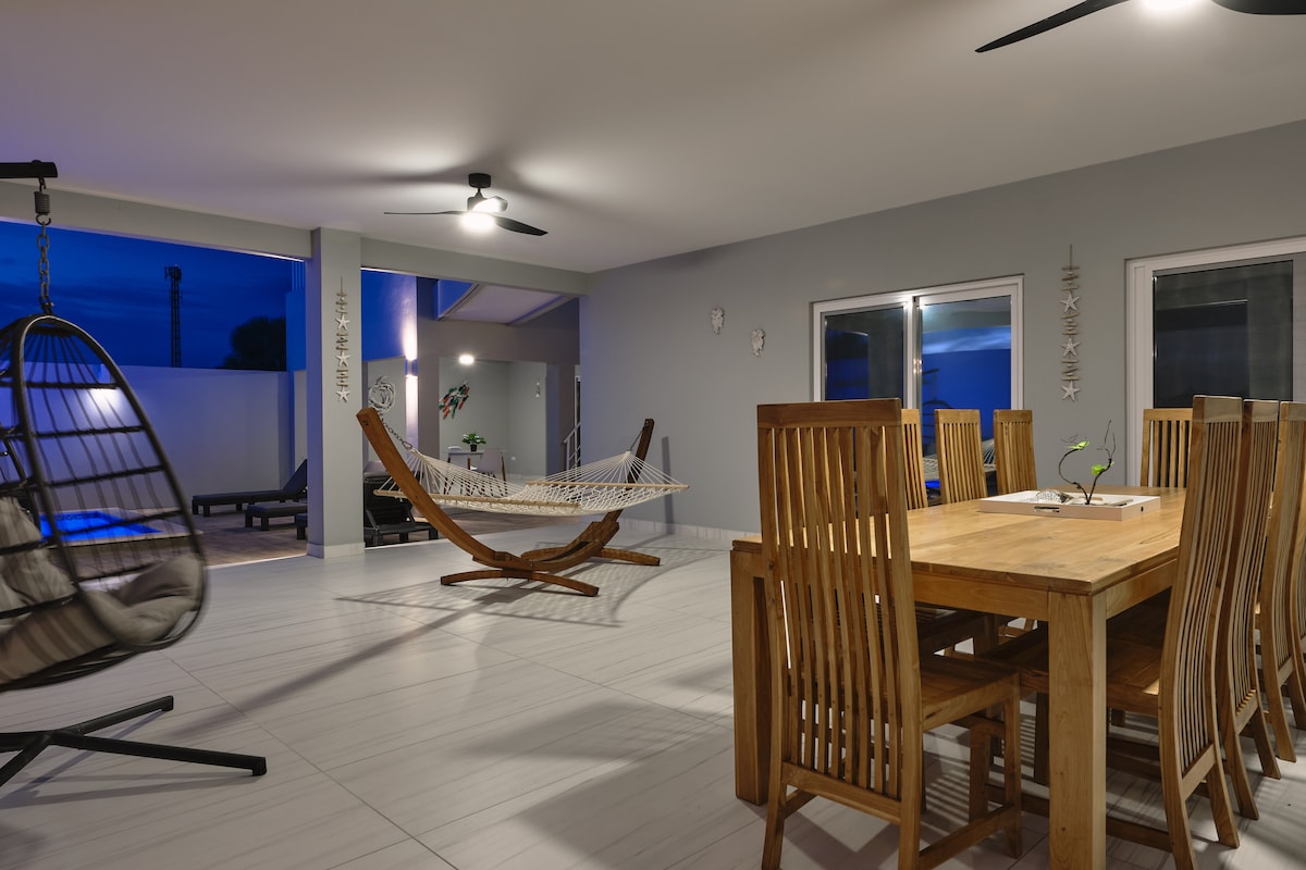 An open-air dining area features a large wooden table surrounded by eight chairs. A hammock and a hanging chair are positioned nearby, providing relaxing seating options. Soft lighting enhances the space, while large windows offer views of the outdoor pool area.