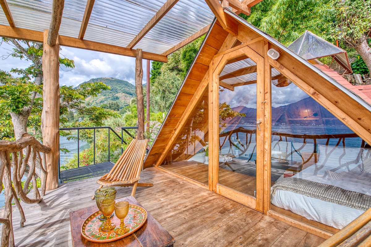 A spacious deck features large windows that provide views of the surrounding mountains and lake. A wooden lounge chair is positioned next to a small table with decorative items. The geometric design of the structure incorporates natural wood elements to blend with the environment.