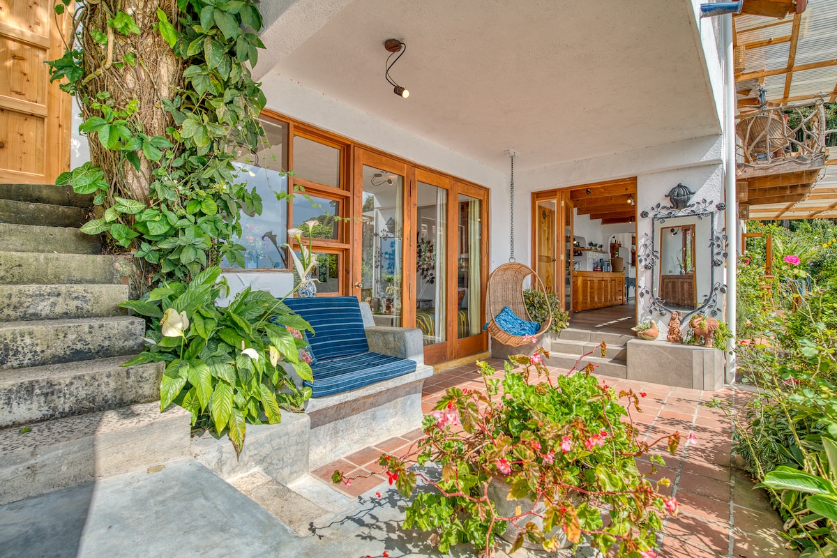 A welcoming entryway features steps leading to a covered porch adorned with potted plants and vibrant flowers. A blue bench and a hanging chair provide seating, while large glass doors invite natural light and views of the indoor space.