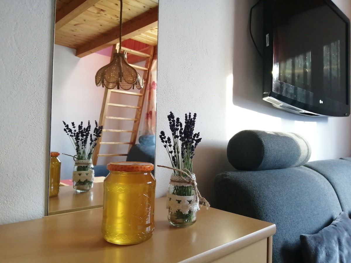 A wooden dresser features a jar of honey and a decorative glass container filled with lavender. A mirror reflects the setting, while a cozy couch and a staircase leading to the upper level are visible in the background. Natural light illuminates the space.