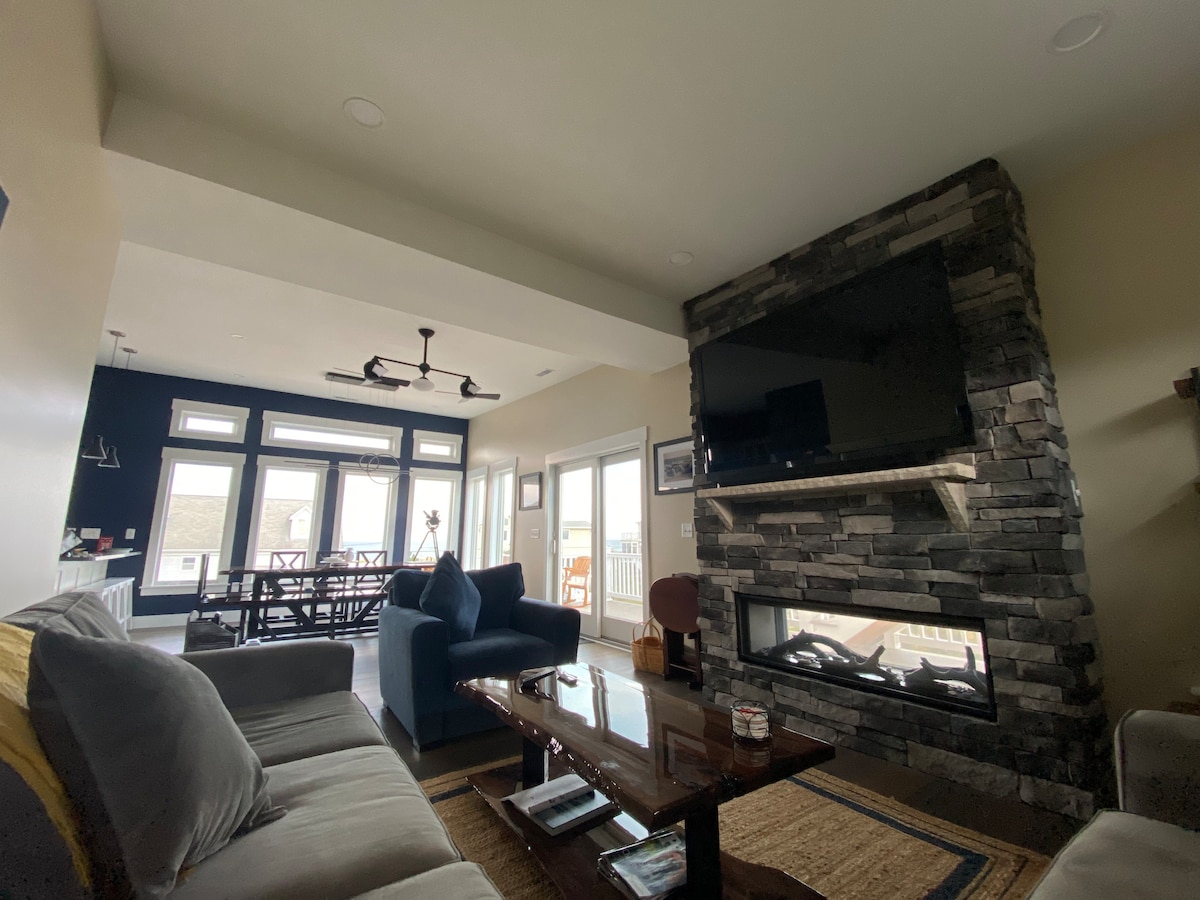 A modern living room features a stone fireplace with a see-thru design, shared with an outdoor deck. Comfortable seating is arranged around a coffee table, and large windows provide ample natural light, overlooking the outdoor space and enhancing the room's spacious feel.