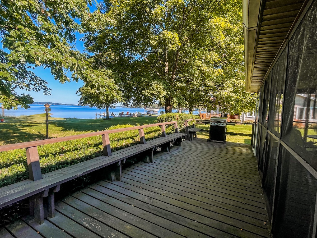 A spacious deck is shown overlooking the well-maintained lawn and Owasco Lake. Adjacent to the deck, a barbecue grill is positioned. Trees provide shade, while the calm water in the distance reflects the clear blue sky.