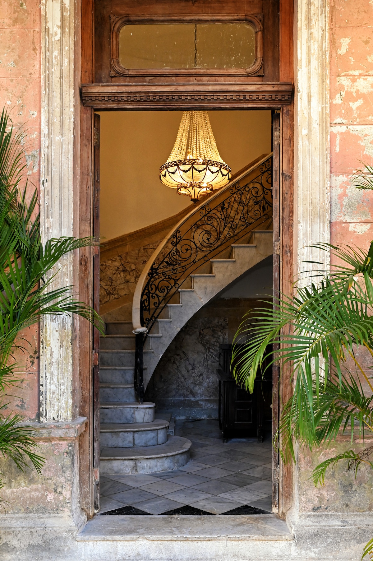 An elegant entrance showcases a vintage chandelier illuminating a winding staircase adorned with intricate ironwork. Potted palms flank the doorway, and light reflects off the polished marble steps, welcoming guests into the hotel’s thoughtfully curated interior.