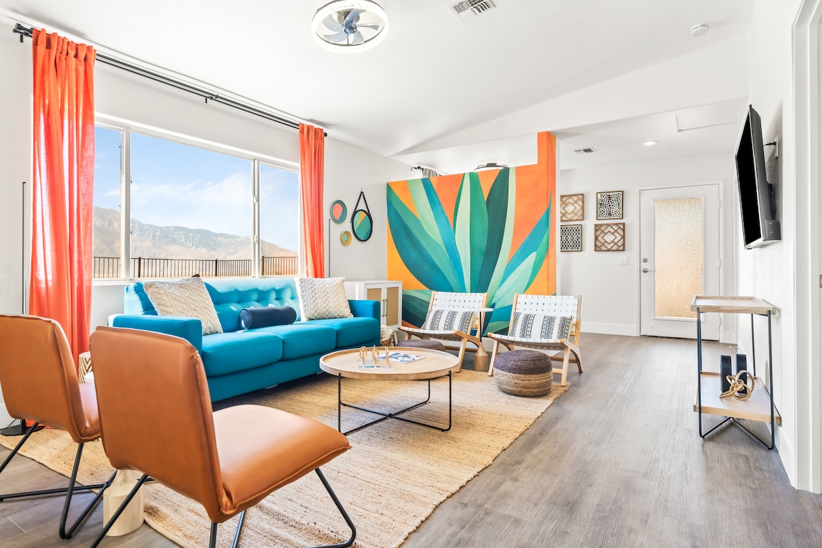 The living area features a vibrant blue sofa paired with two stylish orange chairs, creating a bold contrast. A large window offers natural light and scenic mountain views. Artistic wall decor includes a colorful mural, while a circular coffee table sits atop a textured area rug.