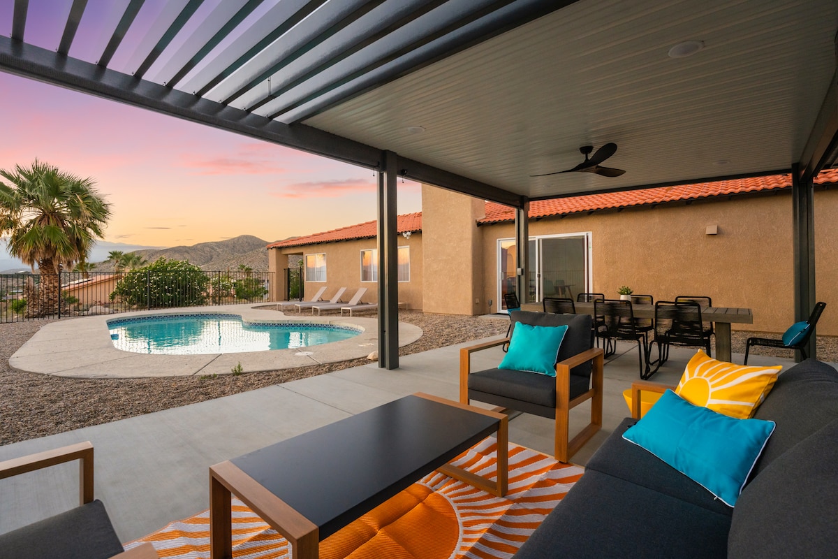 A shaded outdoor seating area features modern lounge furniture with colorful cushions. The private pool glistens in the background, surrounded by desert landscaping. A dining table is positioned nearby, offering an inviting space for meals while enjoying beautiful sunset views.