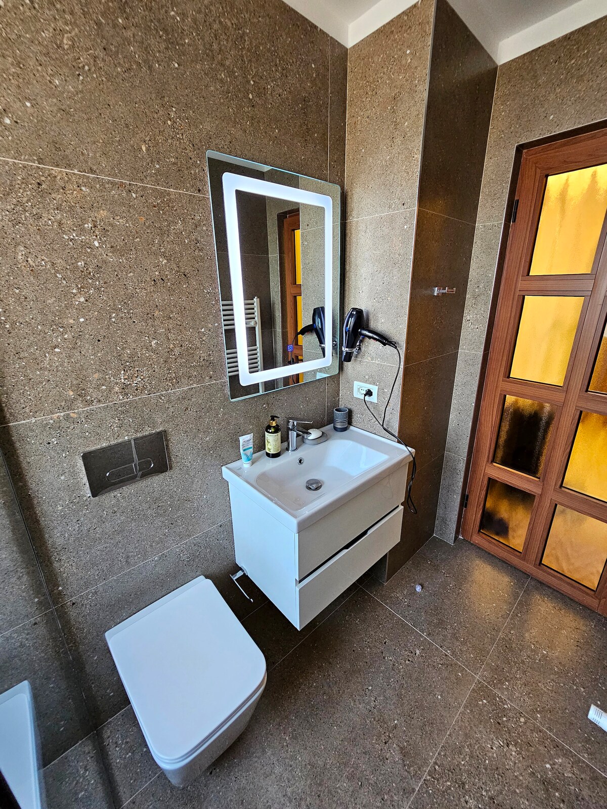 A modern bathroom features stone-textured walls and floor, providing a neutral backdrop. A sleek white vanity with two drawers is positioned below a rectangular mirror, which is illuminated by integrated lighting. A toilet is located beside the vanity, maintaining a clean and functional design.