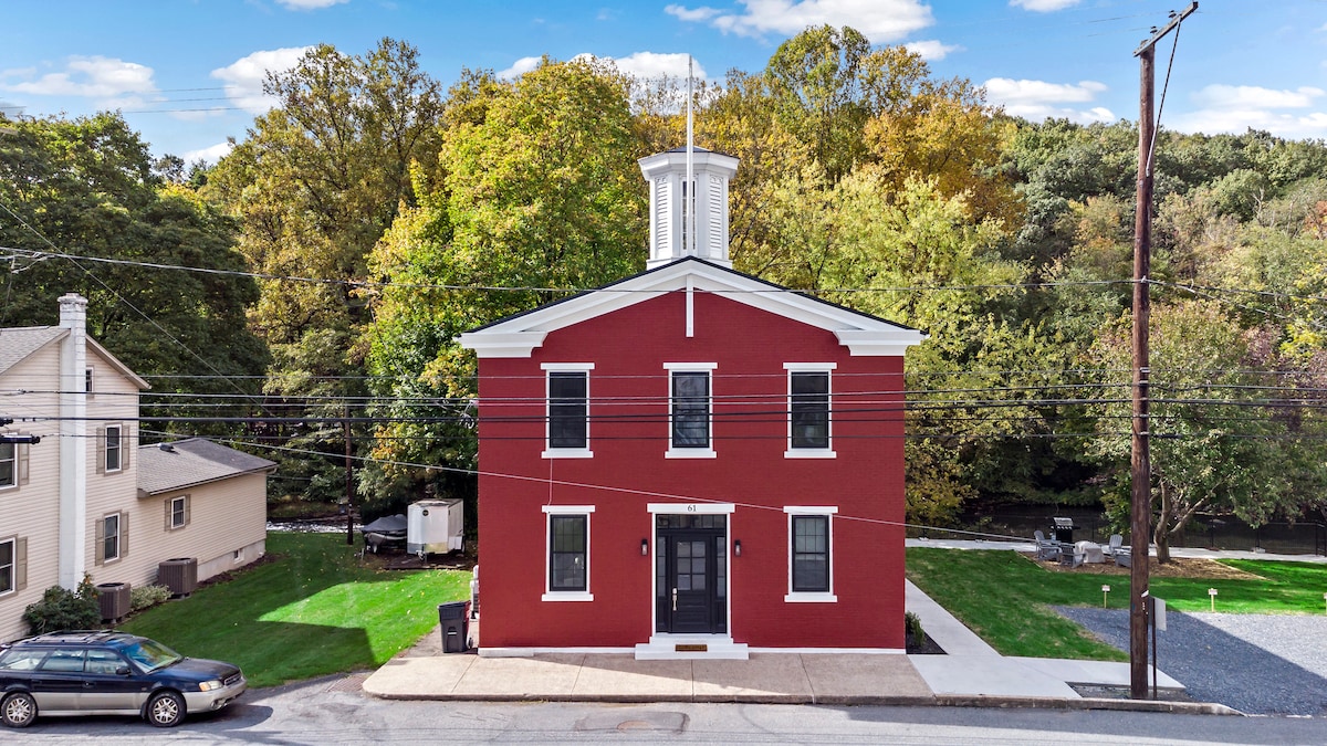 The exterior of a renovated old schoolhouse is showcased, featuring a striking red facade. Surrounded by lush greenery, the building has a classic cupola and evenly positioned windows. A gravel driveway and street parking are visible, enhancing the property’s accessibility.