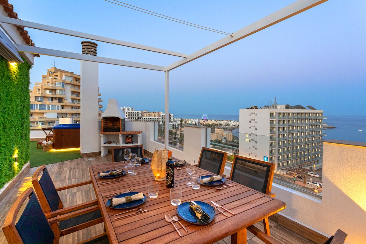 An inviting terrace features a dining table set for four, with views of the sea and surrounding buildings. A barbecue area and a large jacuzzi are also present. Soft lighting creates a welcoming ambiance in the evening sky.