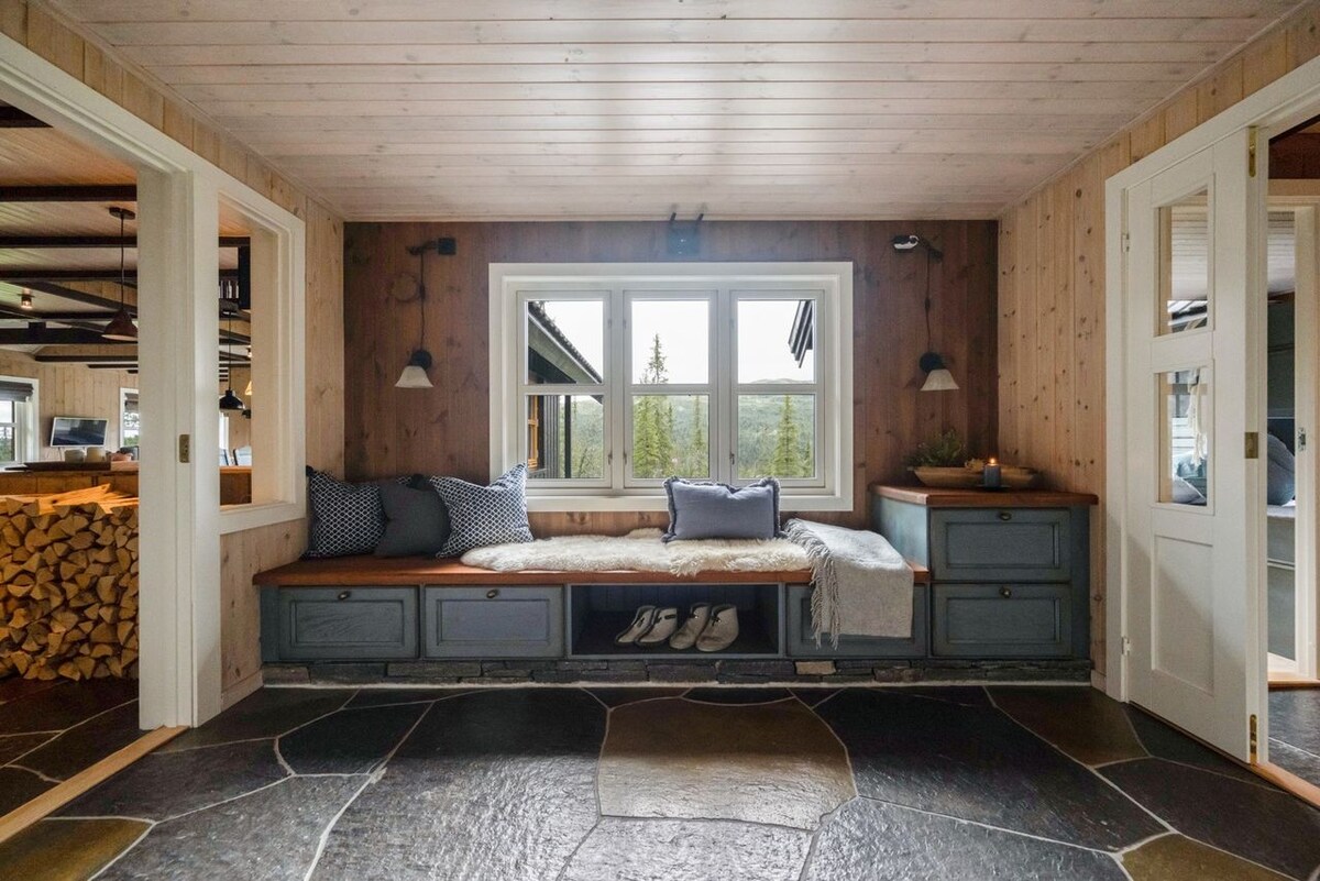 A cozy nook features a built-in bench with plush cushions and a soft blanket, set below large windows. The natural stone floor adds texture to the space, while wooden paneling creates a warm atmosphere.