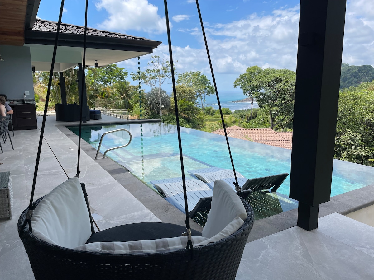 A suspended black wicker chair offers a relaxed spot for overlooking the infinity-edge pool that seamlessly meets the ocean view. Lush greenery surrounds the area, and a shaded patio features sun loungers evocative of a tranquil outdoor space. A glimpse of the coastline is visible in the distance.