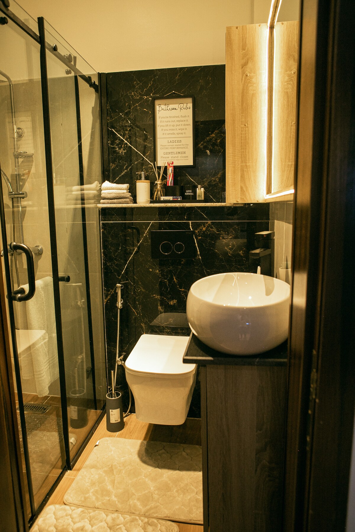 A luxurious bathroom is showcased, featuring a glass shower enclosure and a modern round sink on a wooden counter. Dark marble walls are complemented by soft towels neatly arranged on a shelf. Illuminated mirror enhances the space, creating a sophisticated atmosphere.